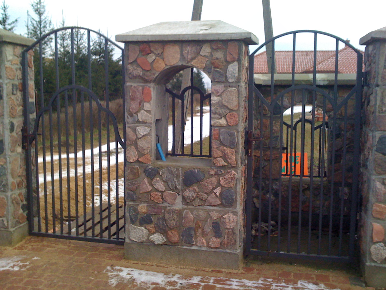 Otwarta brama wjazdowa z czarnego metalu, osadzona w kamiennych słupkach z okienkiem, na ceglanym podjeździe z resztkami śniegu.